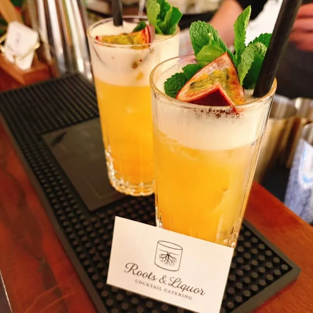 Zwei goldgelbe Cocktails mit Passionsfrucht und frischer Minze neben einer Roots & Liquor Karte.