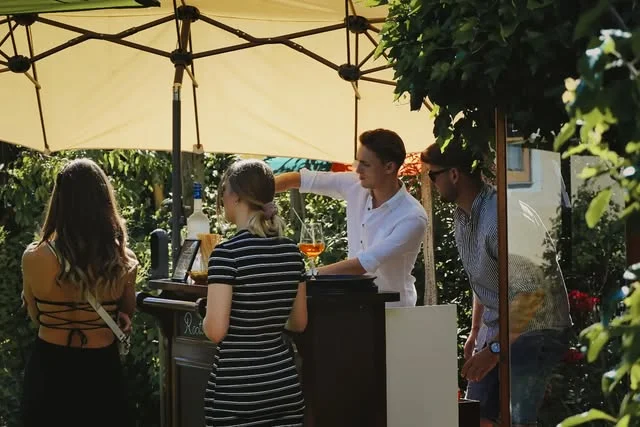 Gäste stehen unter einem Sonnenschirm an der mobilen Roots & Liquor Bar im Garten.